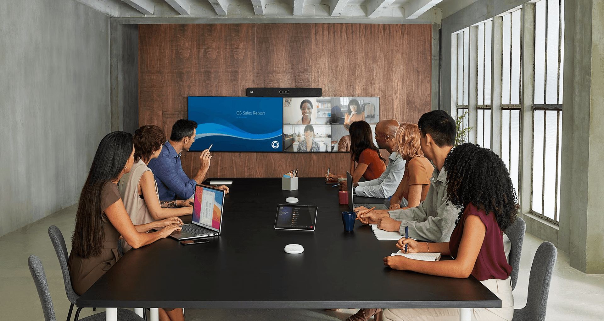 Modern business meeting in a sleek conference room with a black table, professionals using laptops and tablets, and a large screen displaying a Sales Report and video conference participants, highlighting hybrid work and collaboration.